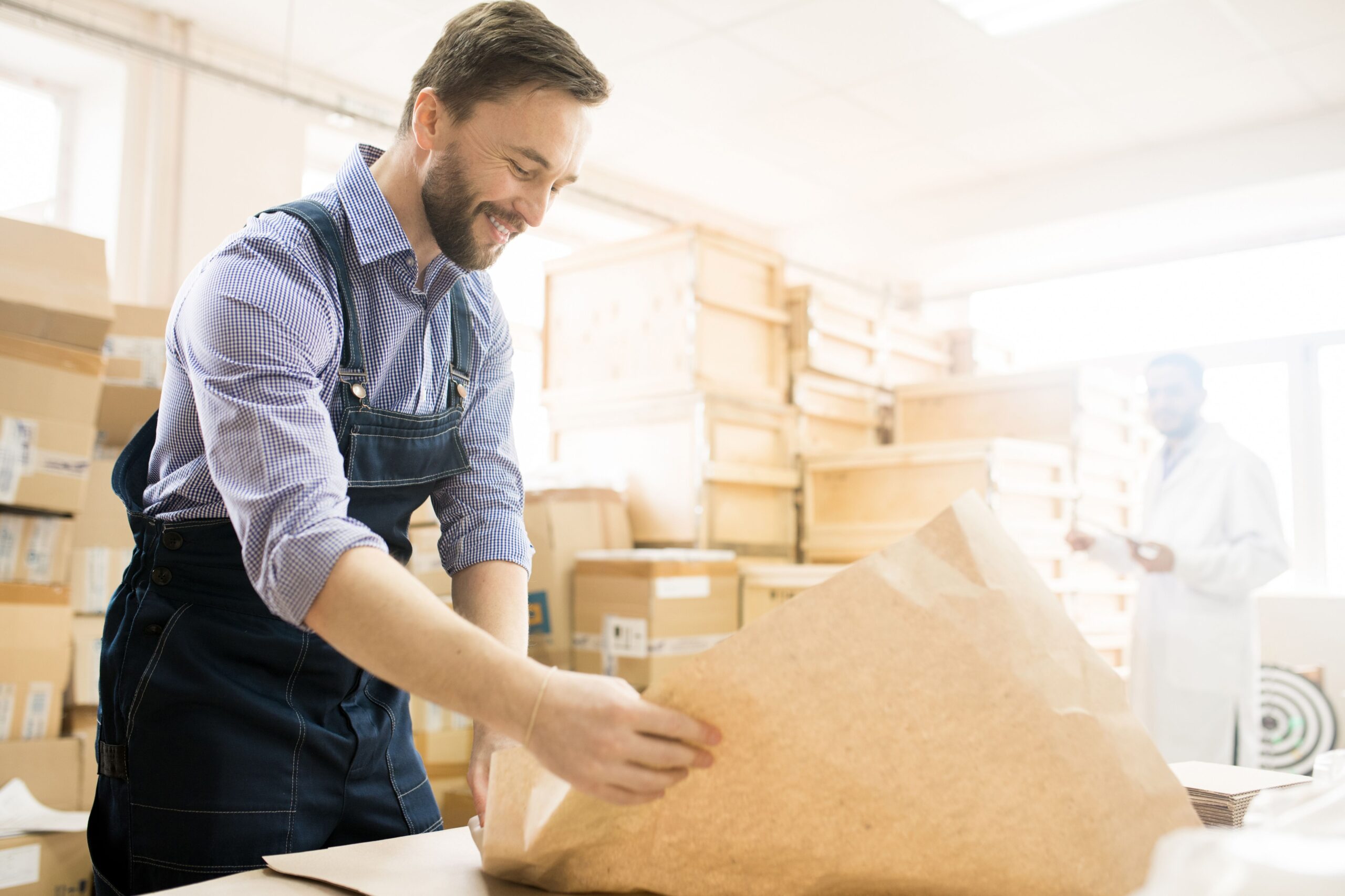Man packing item in brown paper for eco-friendly shipping. Boxes in background.